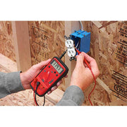 Amprobe 5XP-A compact digital multimeter held in hand while testing a wall electrical outlet with red test leads during wiring work.