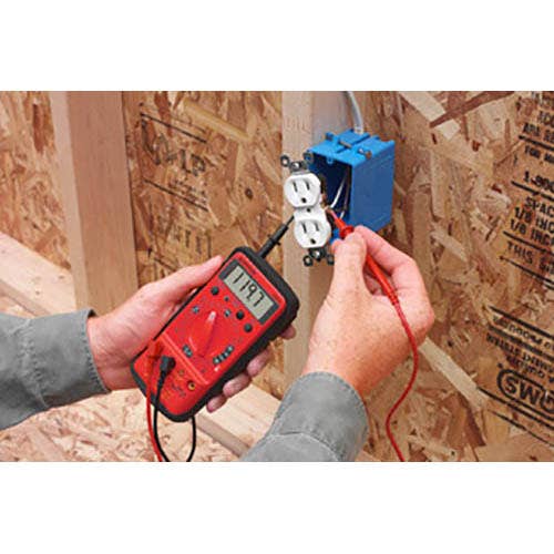 Amprobe 5XP-A compact digital multimeter held in hand while testing a wall electrical outlet with red test leads during wiring work.
